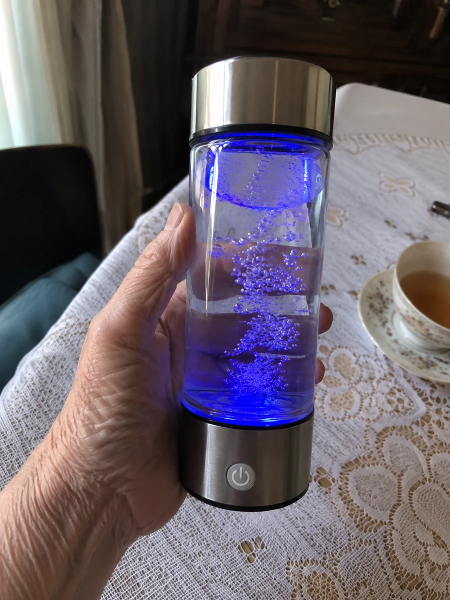 Ръката на възрастна жена държи бутилката Rila Hydrogen с включена синя светлина и водородни мехурчета, на масата до чаша чай